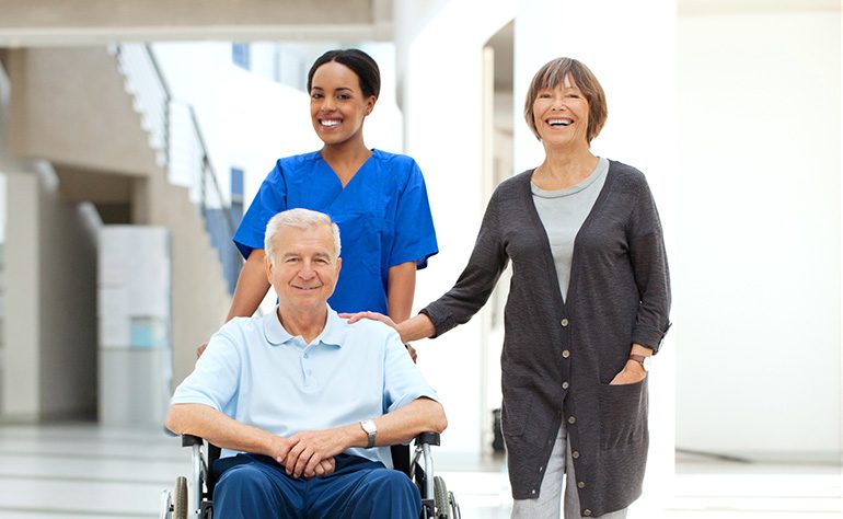 A nurse pushing a smiling man in a wheelchair next to his wife
